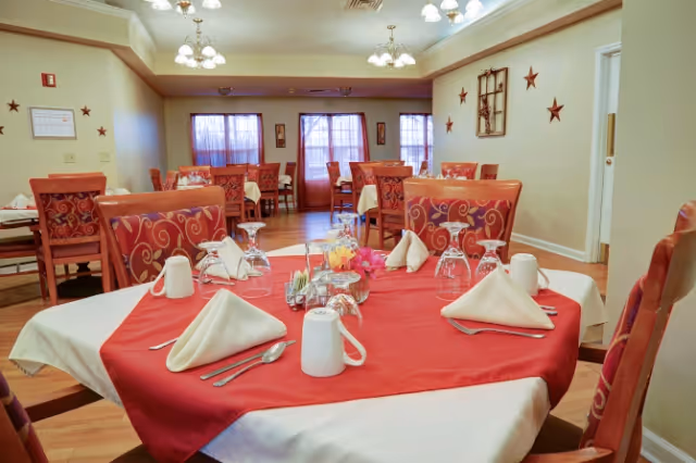 Dining room with multiple tables set with red tablecloths, folded napkins, glassware and chairs in a senior living facility.