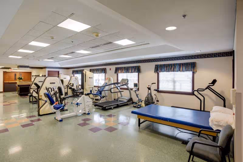 A spacious rehabilitation or physical therapy room with exercise equipment including stationary bikes, treadmills, and weight machines. There is a blue padded therapy table with pillows and a chair nearby. The room has several windows with valances, fluorescent ceiling lights, and a tiled floor with a pattern of purple squares.