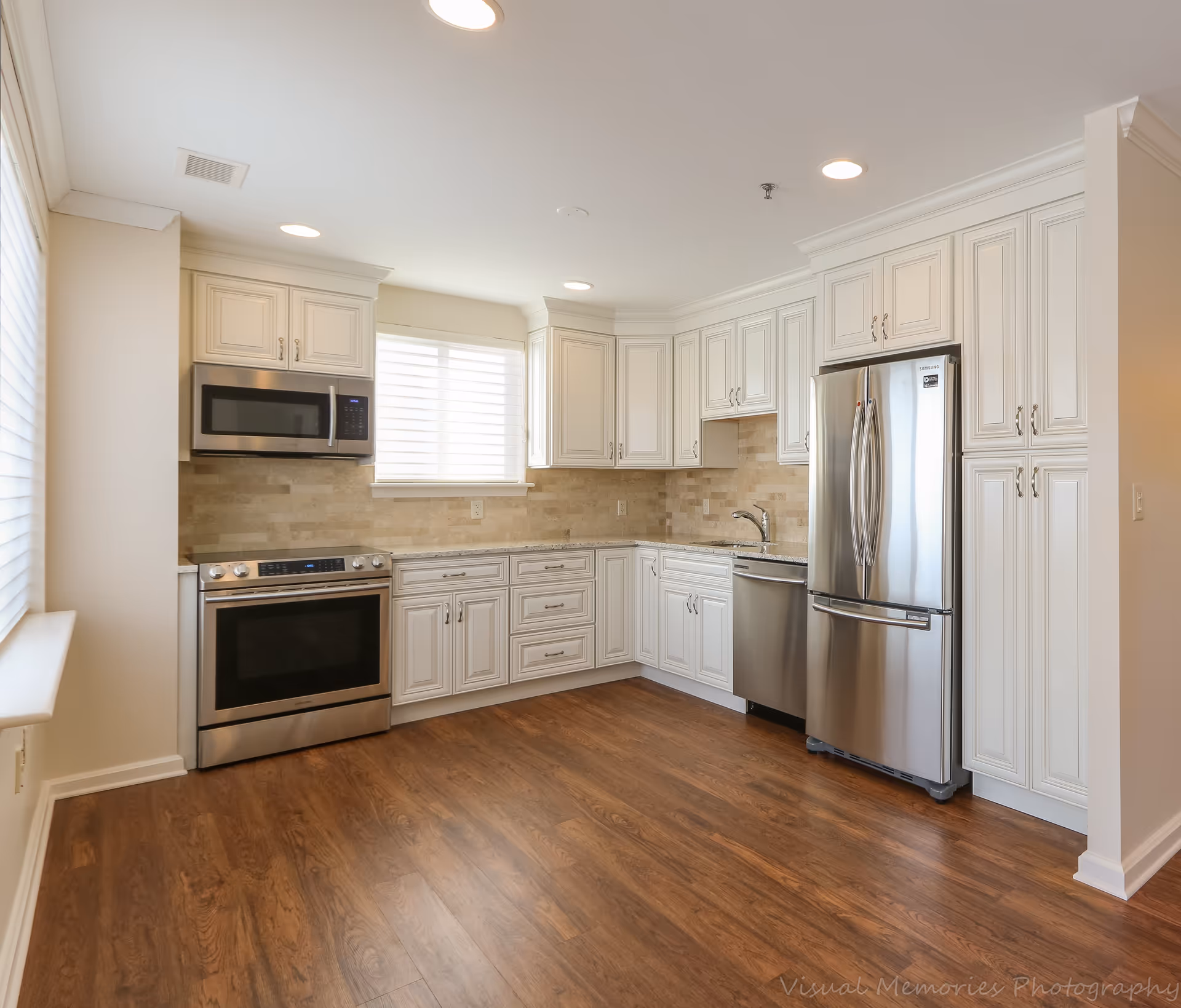 Bright and modern kitchen with white cabinetry, stainless steel appliances including a refrigerator, dishwasher, oven, and microwave. The kitchen has wood flooring, a window with blinds, and recessed ceiling lights.