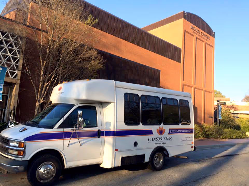 A white shuttle bus with purple and orange stripes parked in front of a brick building labeled 'Welcome Center'. The bus has the Clemson Downs retirement community logo and text on its side. Trees and bushes are visible around the building under a clear blue sky.