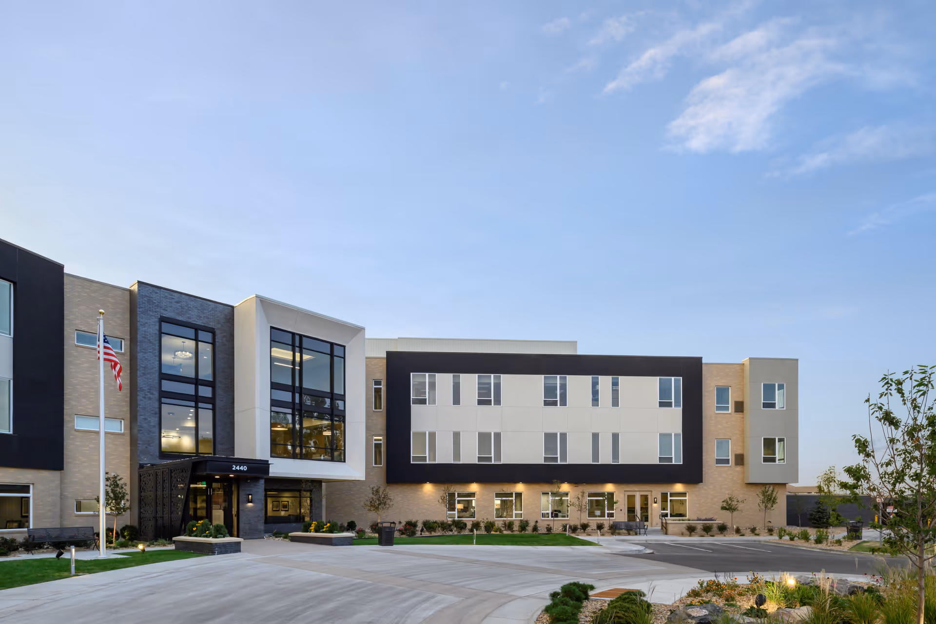 Exterior view of a modern senior living facility building with multiple windows, a flagpole with an American flag, landscaped greenery, and a clear sky.
