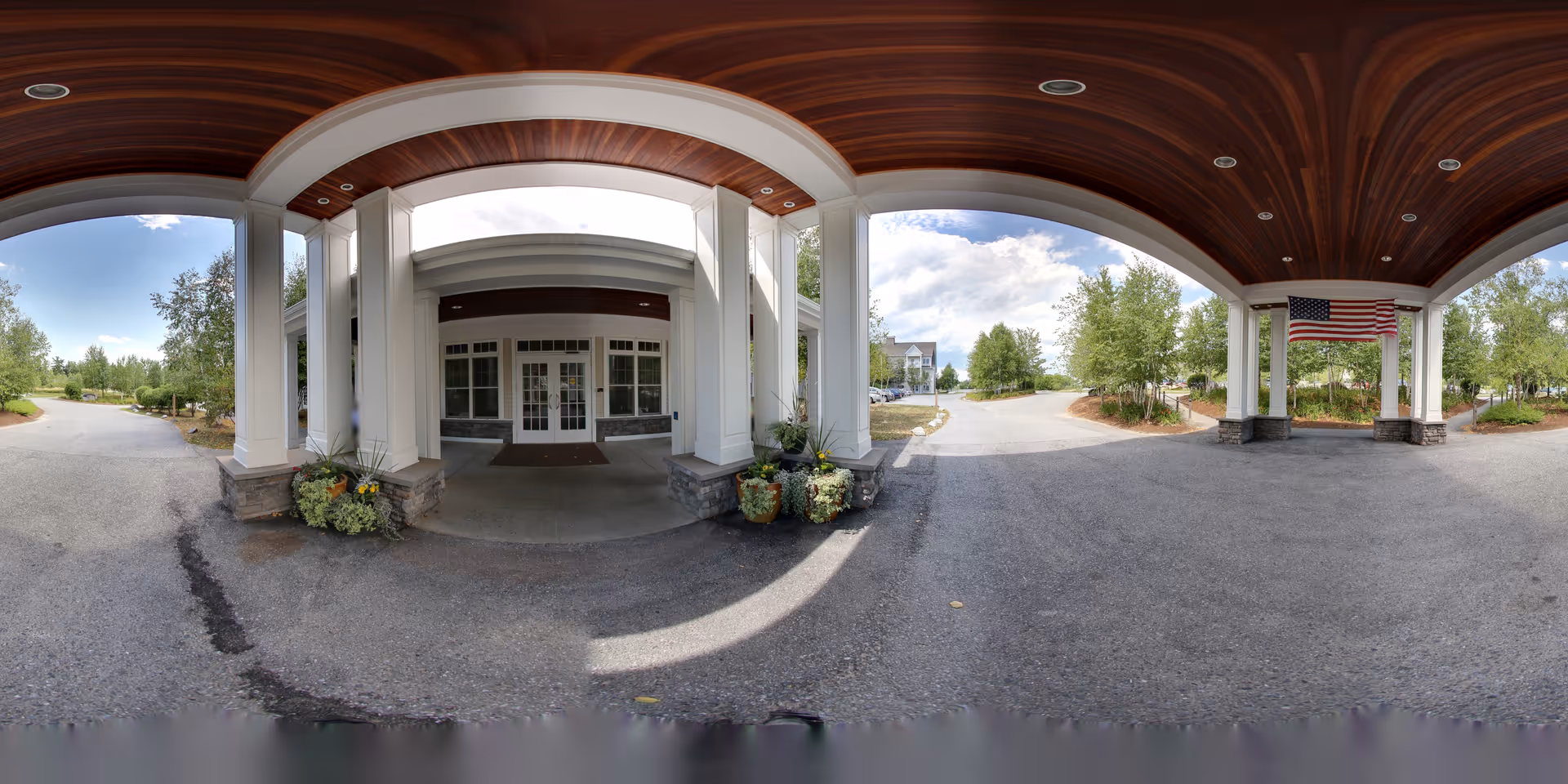 Covered entrance area of The Residence at Otter Creek with white pillars and a wooden ceiling. There are potted plants near the entrance doors, an American flag hanging on the right side, and a driveway leading up to the entrance surrounded by trees and landscaping.