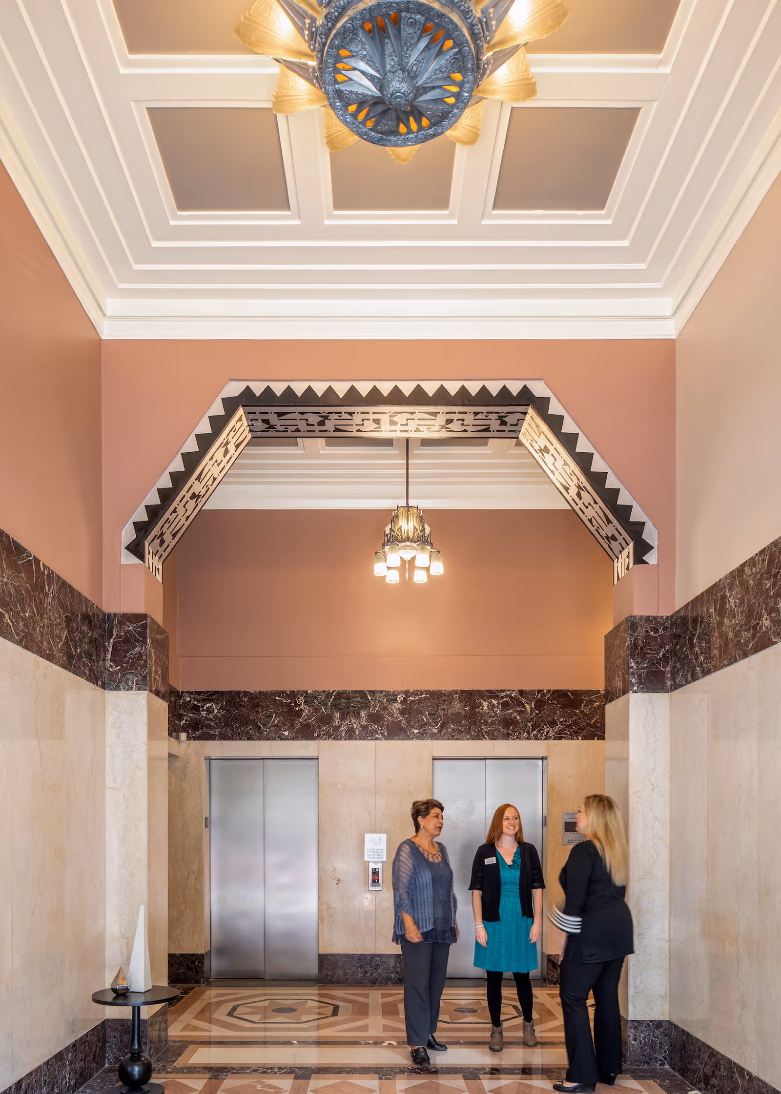 Three women standing and talking in an elegant elevator lobby with marble walls and floors, decorative ceiling, and two closed elevator doors.