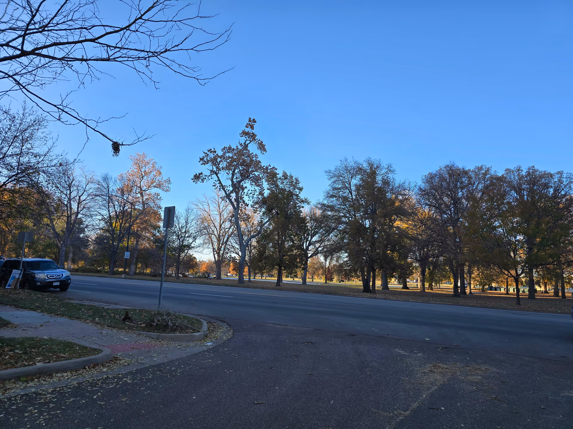A street view showing a road with a parked black SUV on the left side near a sidewalk. Across the road is a park area with many trees, some with autumn-colored leaves. The sky is clear and blue.