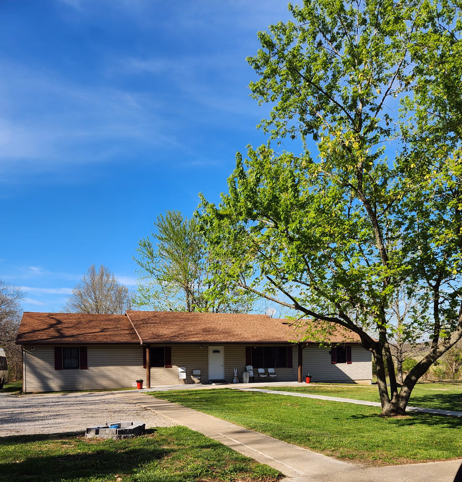 Single-story ranch-style building with a brown roof, front porch seating, a large leafy tree on a green lawn, and a clear blue sky.