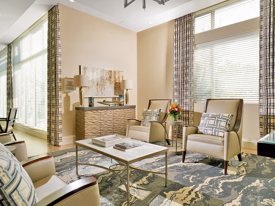 Bright sitting area with upholstered armchairs, a coffee table, sideboard, patterned rug and large windows with blinds.