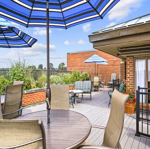 Outdoor patio area with tables and chairs under blue and white striped umbrellas, surrounded by brick walls and greenery, under a partly cloudy sky.