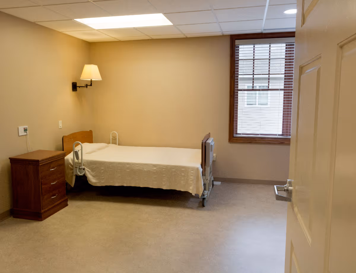 A sparse senior-care bedroom with a hospital-style bed, wooden nightstand, wall lamp, and a window with blinds.