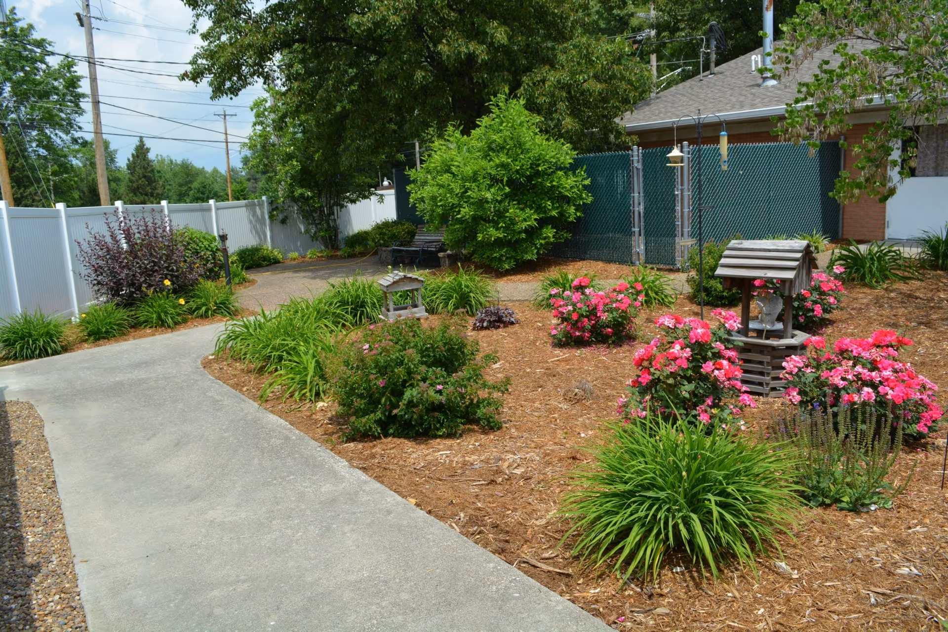 A landscaped outdoor garden courtyard with a concrete walkway, flowering shrubs, and a small decorative wooden well.