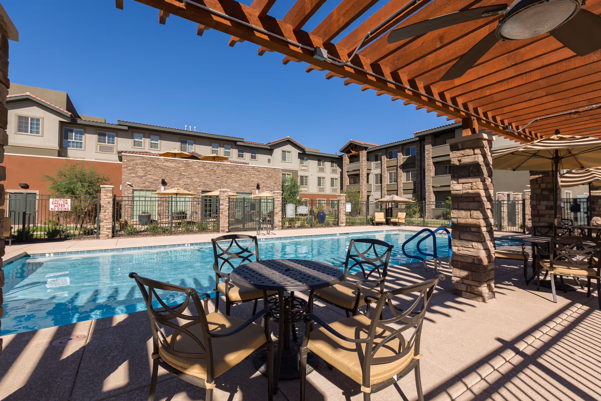 Outdoor swimming pool area at The Enclave at Anthem Senior Living with a round table and four chairs under a wooden pergola. The pool is surrounded by a fence, and there are multi-story residential buildings in the background under a clear blue sky.
