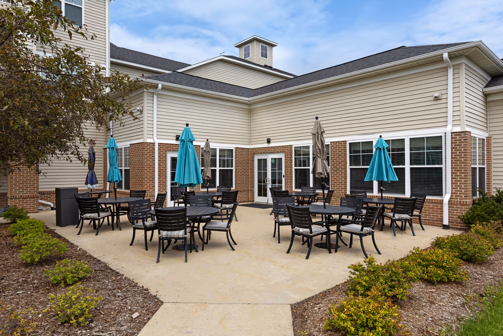 Outdoor patio area at StoryPoint Portage with multiple round tables and black chairs, some tables shaded by blue and beige umbrellas. The patio is surrounded by low bushes and landscaping, adjacent to a beige and brick building under a partly cloudy sky.