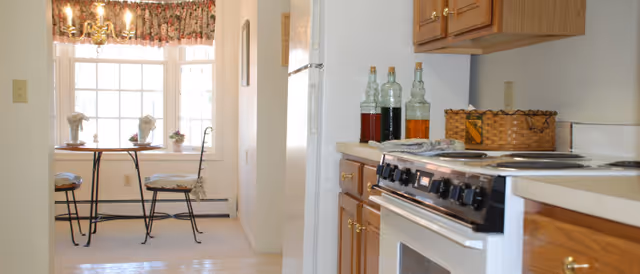 A bright kitchen area with wooden cabinets, a white stove, and three decorative bottles on the counter. In the background, there is a small dining nook with a round table, two chairs with cushions, and a large window with floral curtains letting in natural light.