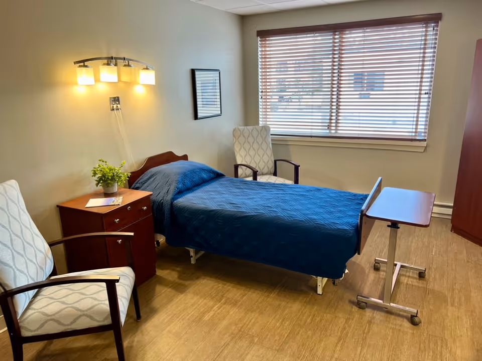 A small, neatly arranged bedroom in a skilled nursing facility with a single bed covered in a blue quilt, two patterned armchairs, a wooden nightstand with a small plant and papers, a wall-mounted light fixture above the bed, a window with wooden blinds, and a mobile overbed table on wheels.