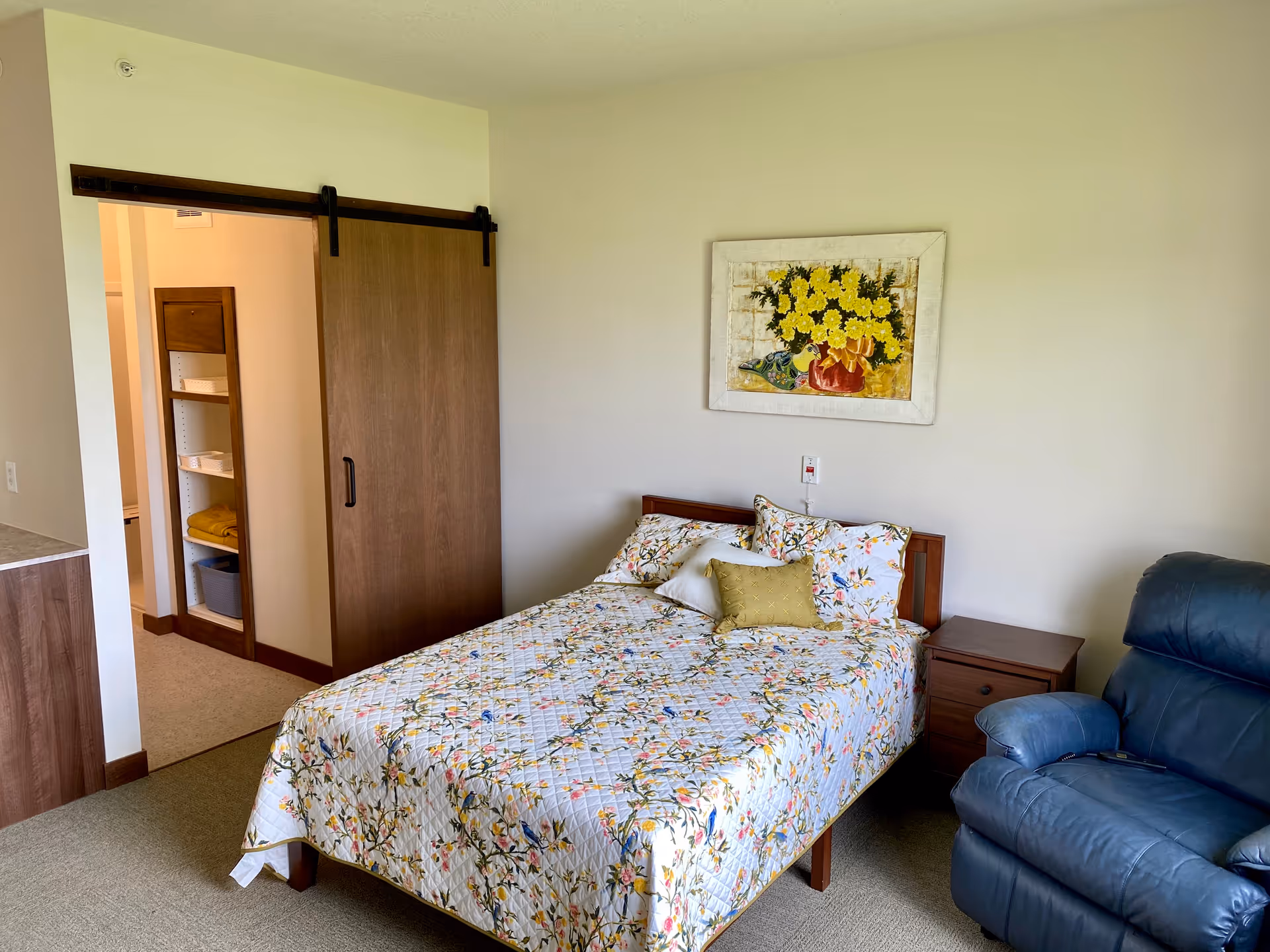 A furnished bedroom with a floral quilted bed, wooden nightstand, blue recliner, and a sliding barn-door closet.