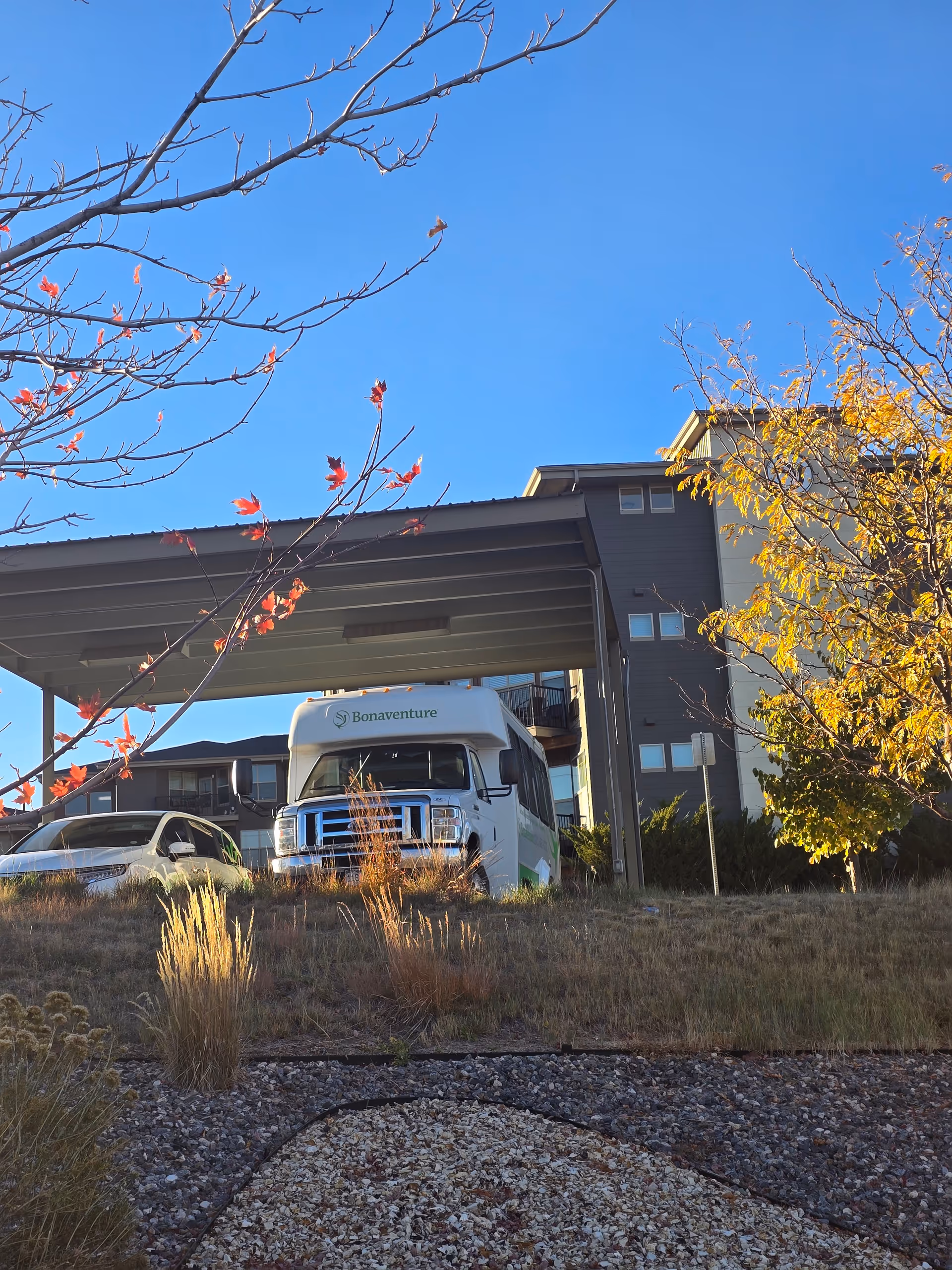 A white Bonaventure shuttle bus and a white car parked under a large covered area in front of a multi-story building. The scene is outdoors with dry grass, some bushes, and trees with autumn-colored leaves under a clear blue sky.
