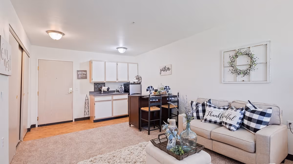 A cozy senior living apartment interior featuring a small kitchen area with white cabinets and a countertop, a dining table with two chairs, and a beige sofa adorned with black and white checkered pillows and a pillow that says 'Always be Thankful'. The walls are decorated with a wreath and signs that say 'gather' and 'Welcome friends'. The floor is a combination of carpet and wood.