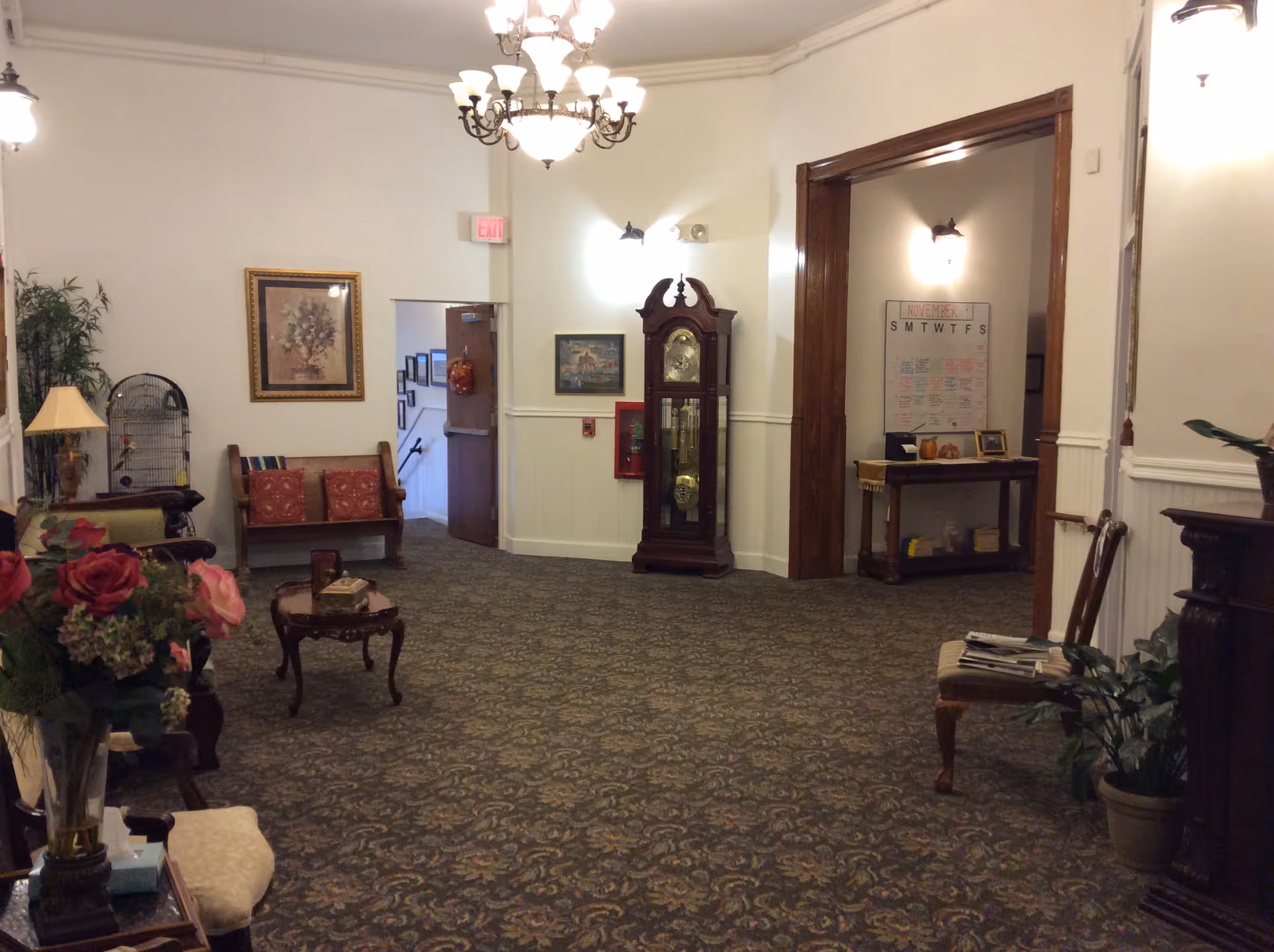 Interior view of a senior living facility common area with patterned carpet, antique-style furniture including chairs, a small table, a grandfather clock, framed artwork on the walls, potted plants, and a chandelier hanging from the ceiling.