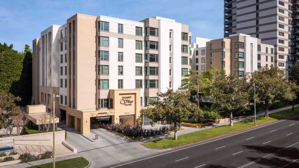 Exterior view of Belmont Village Senior Living Westwood, a multi-story beige and white building with multiple windows, surrounded by trees and greenery, located next to a street with a sidewalk.