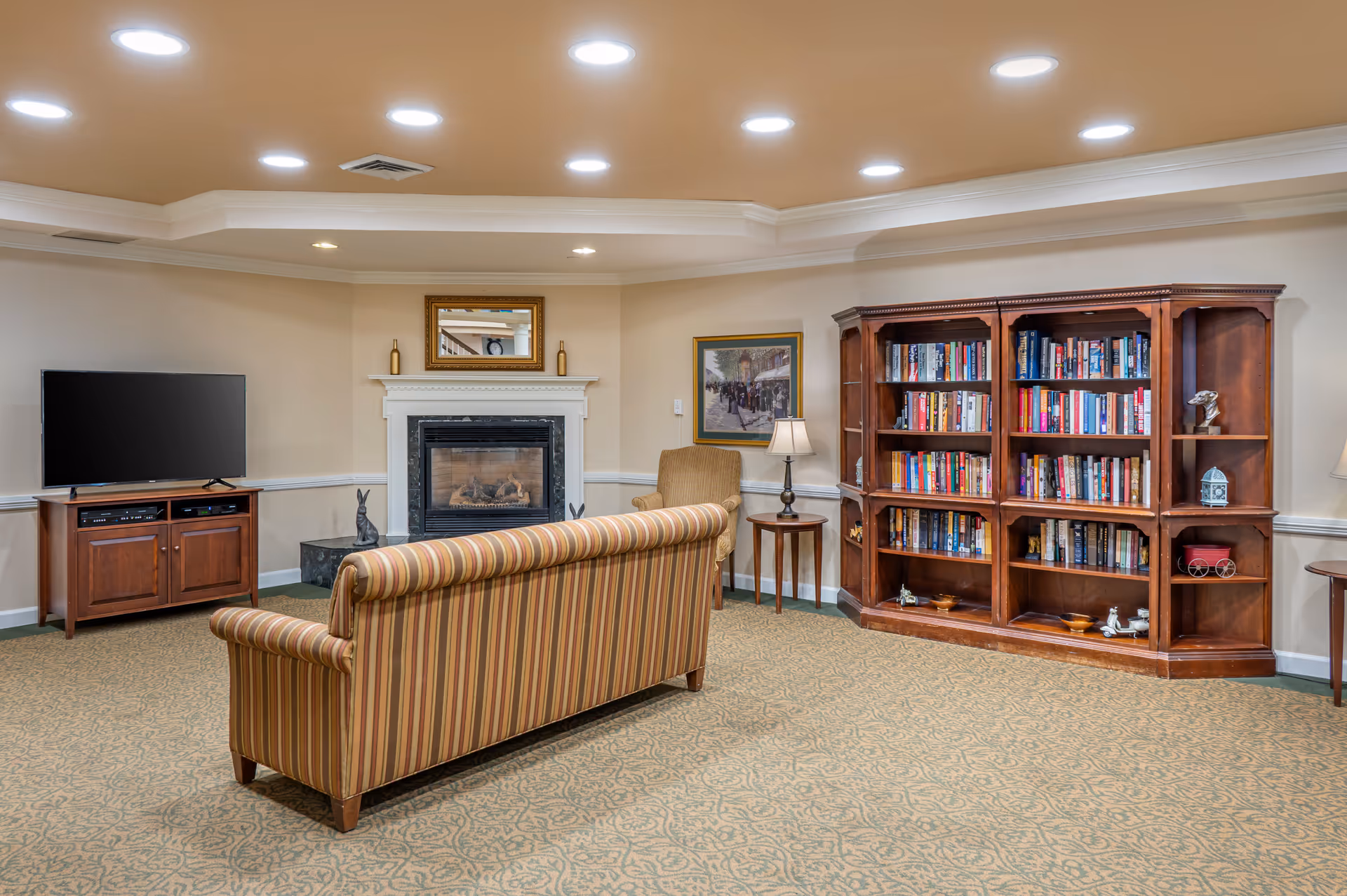A cozy living room area with a striped sofa facing a fireplace and a flat-screen TV on a wooden stand. To the right, there is a large wooden bookshelf filled with books and decorative items. A cushioned armchair and a small side table with a lamp are positioned near the fireplace. The room has beige walls, a patterned carpet, and recessed ceiling lights.