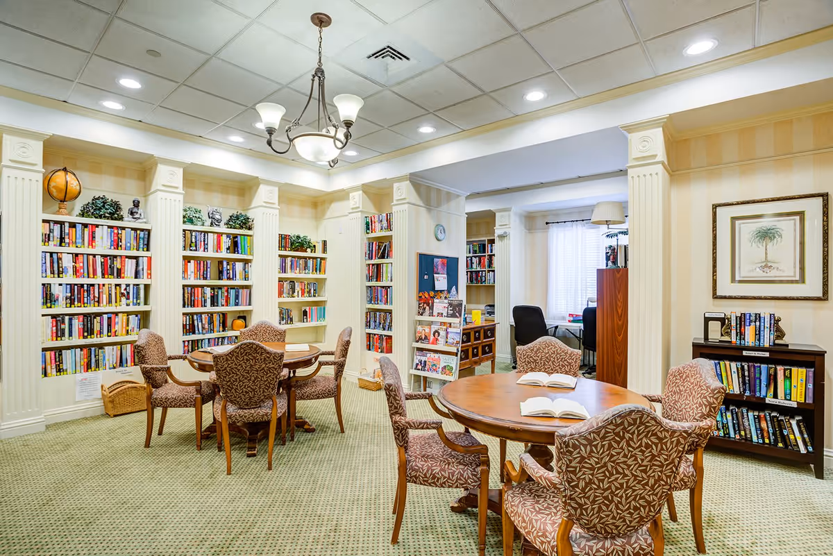 A well-lit library room with multiple bookshelves filled with books and decorative plants. There are round wooden tables with patterned upholstered chairs around them. The room has beige walls with white columns and a chandelier hanging from the ceiling. In the background, there is a small office area with a desk and chairs near a window with sheer curtains.