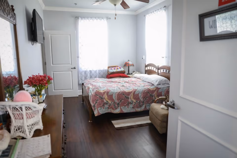 Sunlit bedroom with a floral-patterned bed, dresser topped with flowers and decor, a wall-mounted TV, and an armchair.