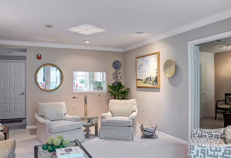A cozy living room with two cushioned armchairs and a patterned sofa. The walls are decorated with a round mirror, three decorative plates, a framed landscape painting, and a large decorative plate. A small side table with a lamp is positioned between the armchairs. A glass coffee table with books and decorative green spheres is in the foreground. The room has light-colored walls and carpet, with a doorway leading to another room visible on the right.