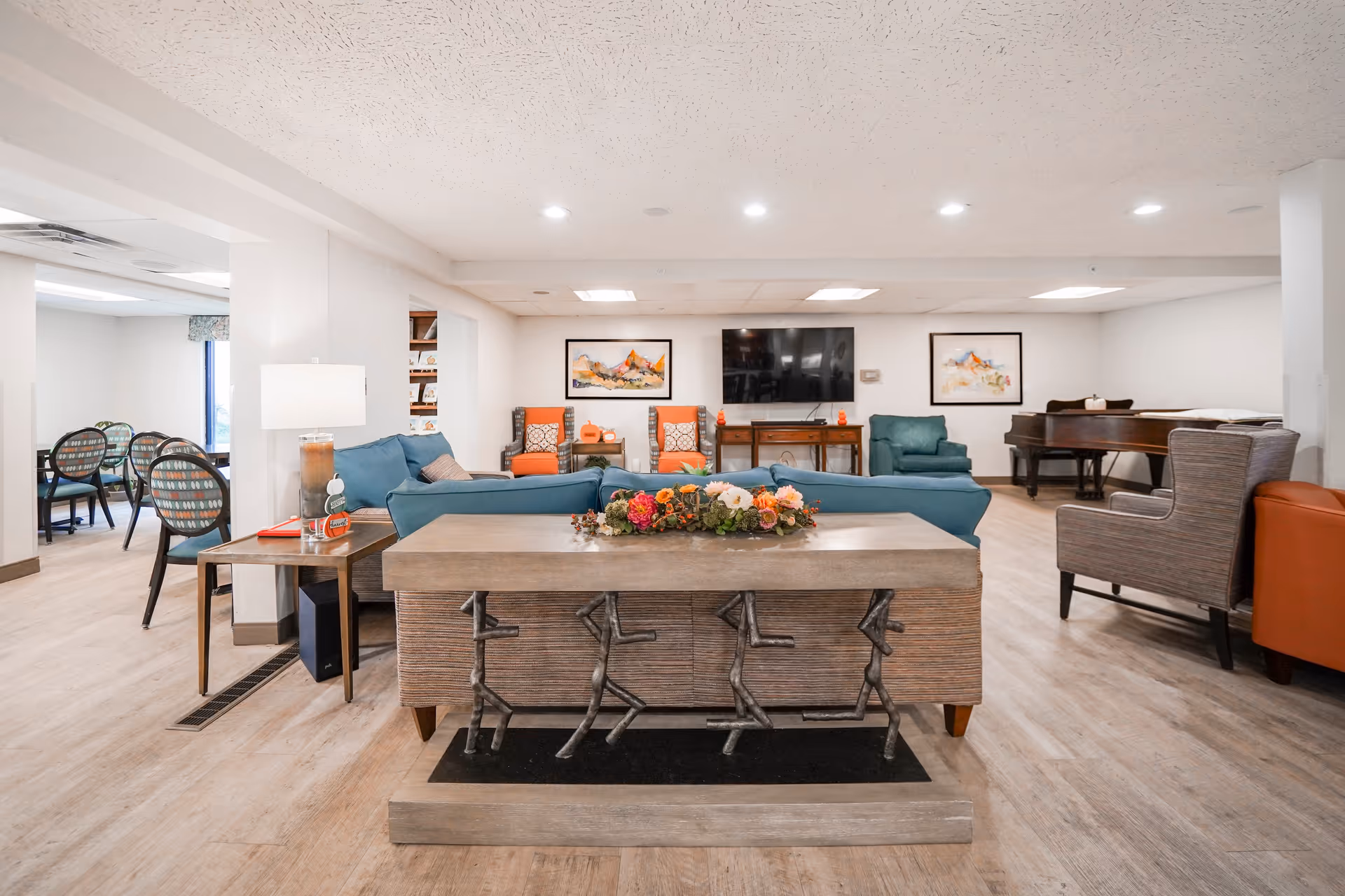 A bright senior living common room with teal sofas, armchairs, a TV on the far wall, a piano, and a dining area to the left.
