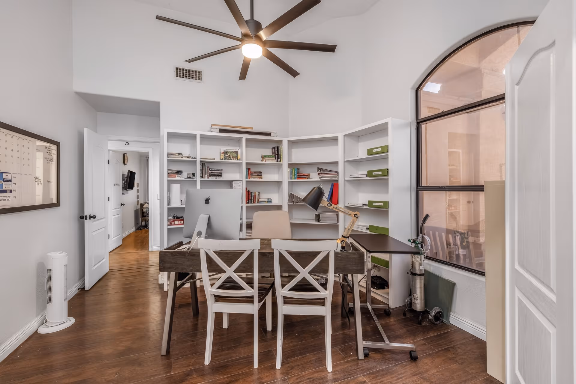 A bright office room with wooden flooring, a large ceiling fan, and a window with an arched top. The room contains a desk with two white chairs, a computer, a desk lamp, and bookshelves filled with books and files. A whiteboard calendar is mounted on the wall to the left, and an open door leads to another room.