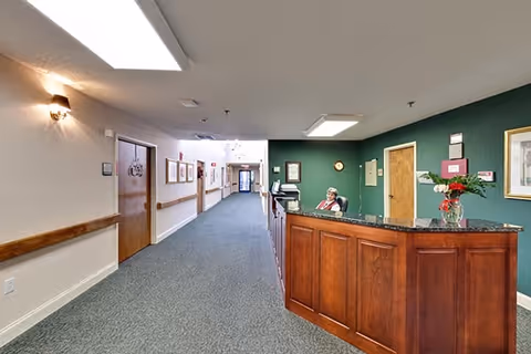 Interior view of a senior living facility hallway with a reception desk on the right side. A staff member is seated behind the wooden reception desk with a granite countertop. The walls are painted green behind the desk and beige along the hallway, which is carpeted in blue. Several doors and framed pictures line the hallway, and ceiling lights illuminate the space.