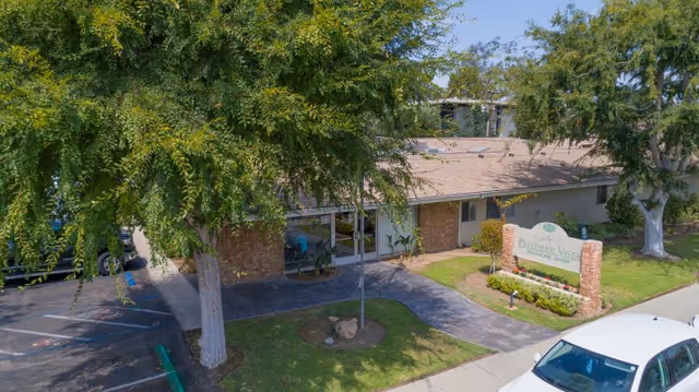 Front exterior of a single-story healthcare building with trees, a parking area, walkway, and a sign for Palomar Vista Healthcare Center.