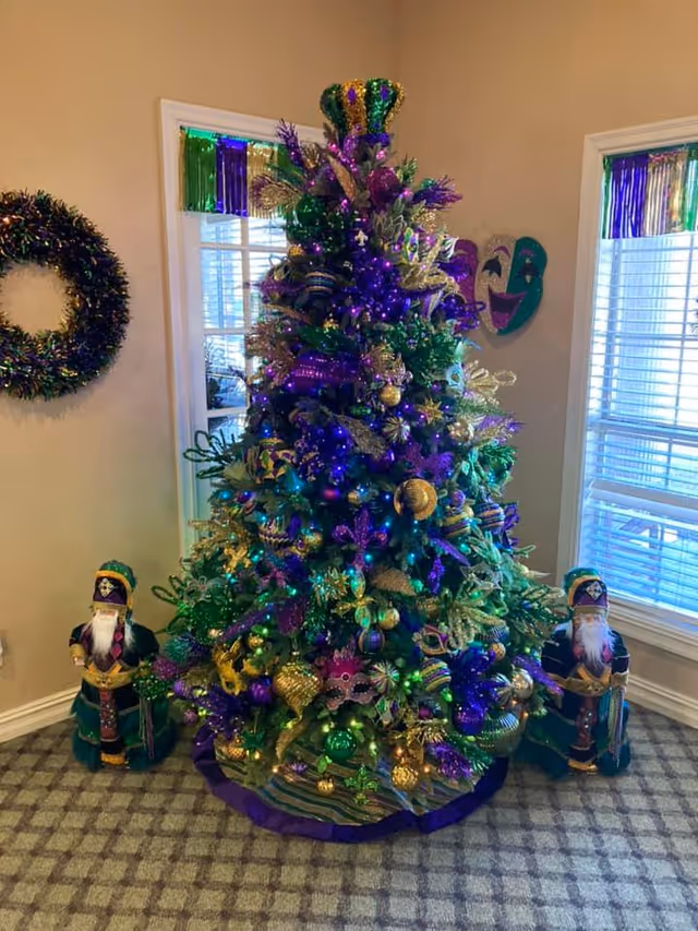A Christmas tree decorated in purple, green, and gold ornaments with Mardi Gras themed decorations including masks and beads, topped with a crown. The tree is placed in a corner of a room with two windows behind it, each adorned with purple, green, and gold fringe decorations. There are two nutcracker figures dressed in matching Mardi Gras colors on either side of the tree, and a wreath decorated in similar colors hangs on the wall to the left.