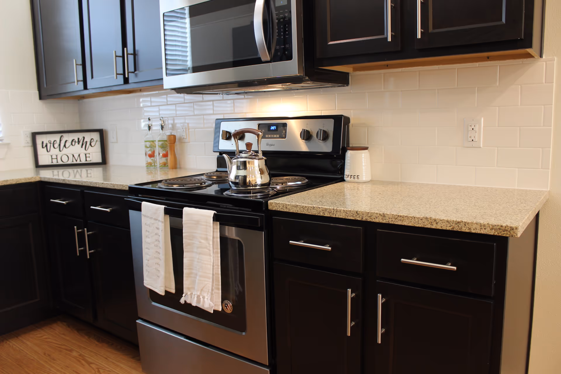 Modern kitchen with dark cabinets, granite countertops, a stainless steel stove and microwave, a kettle on the range, and a 'Welcome Home' sign on the counter.