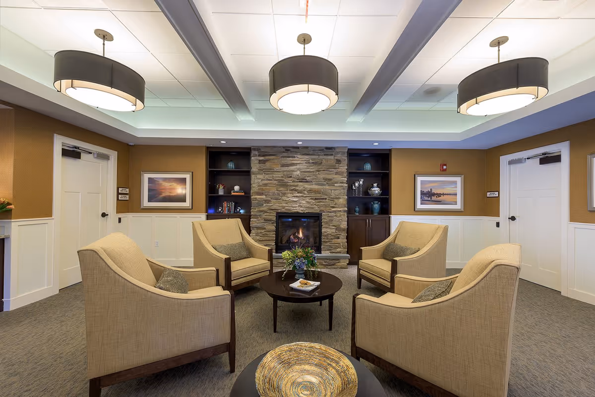 A cozy seating area in a senior living facility with four beige armchairs arranged around a round wooden coffee table. The room features a stone fireplace in the center with built-in shelves on either side displaying decorative items. The walls are painted in warm tones with white wainscoting, and there are two framed landscape pictures on the walls. The ceiling has recessed lighting and three large modern pendant lights.