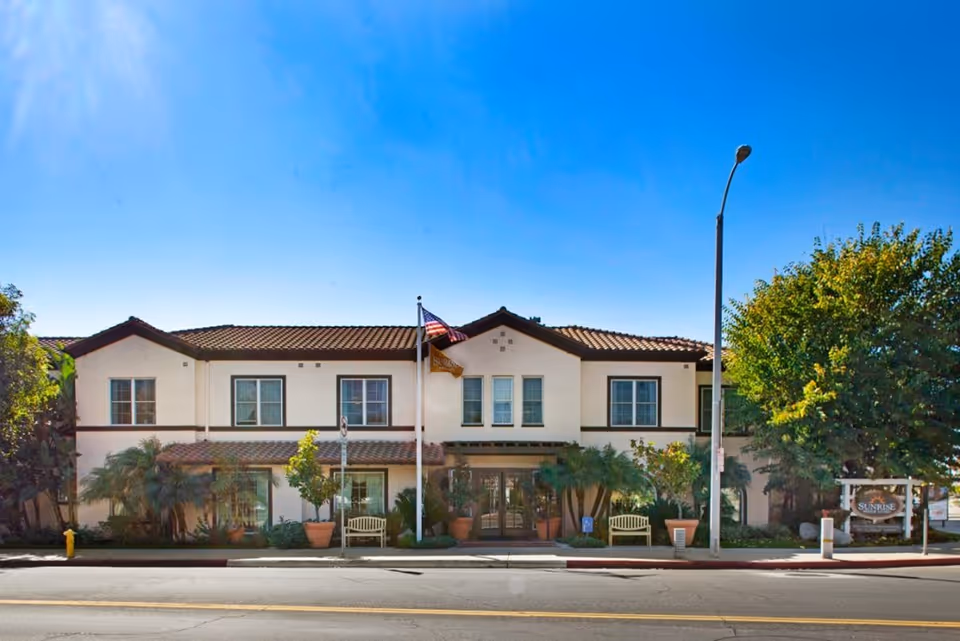 Front exterior of a two-story stucco senior living building with a tiled roof, American flag, benches and landscaped plants.
