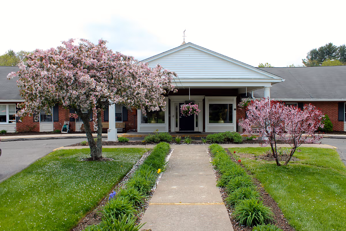 Front entrance of a one-story brick facility with a covered portico, a concrete walkway, and blooming pink trees in the landscaped lawn.