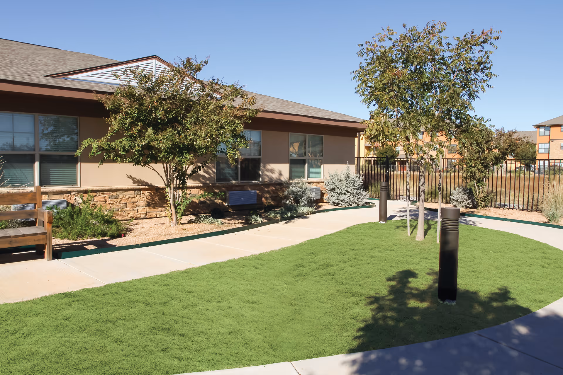 Outdoor courtyard area at The Courtyards Assisted Living and Memory Care featuring a curved concrete walkway, green grass, small trees, shrubs, a wooden bench, and a building with windows in the background under a clear blue sky.