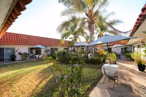 Sunny courtyard of a senior living facility with tables, chairs, umbrellas, and palm trees.