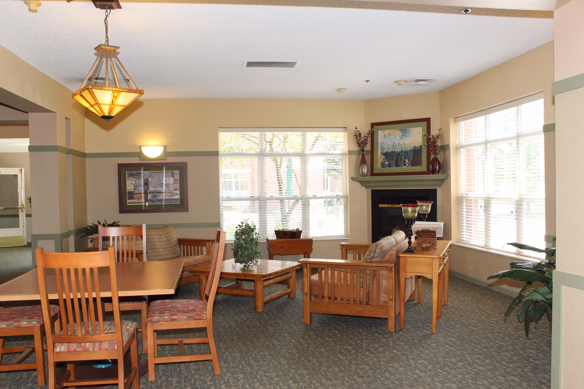 A cozy living room area in Main Street Lodge with wooden furniture including a dining table with chairs, a coffee table, and two armchairs. The room features large windows with blinds allowing natural light to fill the space. There is a fireplace with a painting above it and decorative vases on the mantel. The walls are painted beige with green trim, and the carpet is patterned. A hanging light fixture is visible above the dining table.