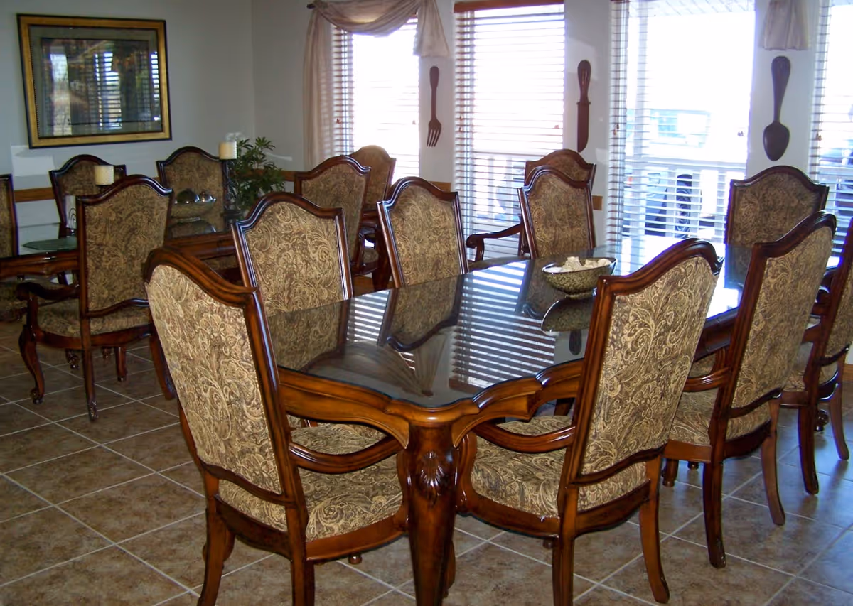A formal dining room with a large rectangular glass-top wooden table surrounded by eight upholstered chairs with ornate wooden frames. The room has tiled flooring, large windows with blinds, and decorative wall hangings shaped like a fork and spoon. There is a framed picture on one wall and a small plant on a side table in the background.