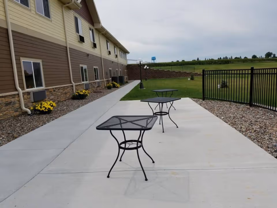 Concrete patio with three metal mesh tables beside a senior living building and a fenced grassy lawn.