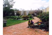 Brick courtyard path leading through a landscaped garden with potted flowers and low building walls under a partly cloudy sky.