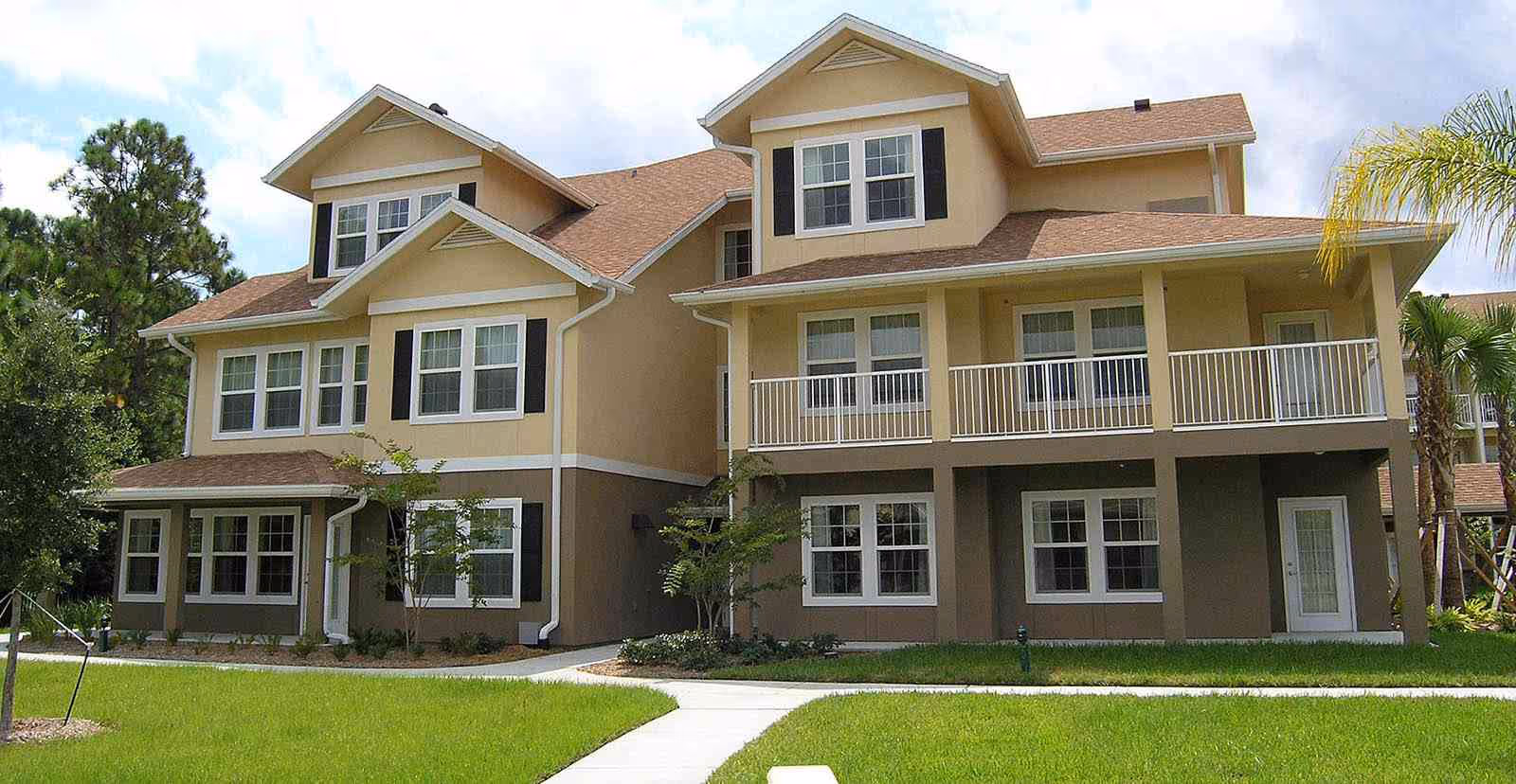 Exterior view of a two-story residential building with beige and brown walls, multiple windows with white frames, and a balcony on the second floor. The building is surrounded by a well-maintained lawn, small trees, and a concrete walkway leading to the entrance. The sky is partly cloudy.