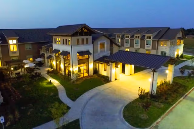 Evening aerial view of The Healthcare Resort of Plano building with exterior lights on, showing a covered entrance driveway, landscaped pathways, and surrounding greenery.