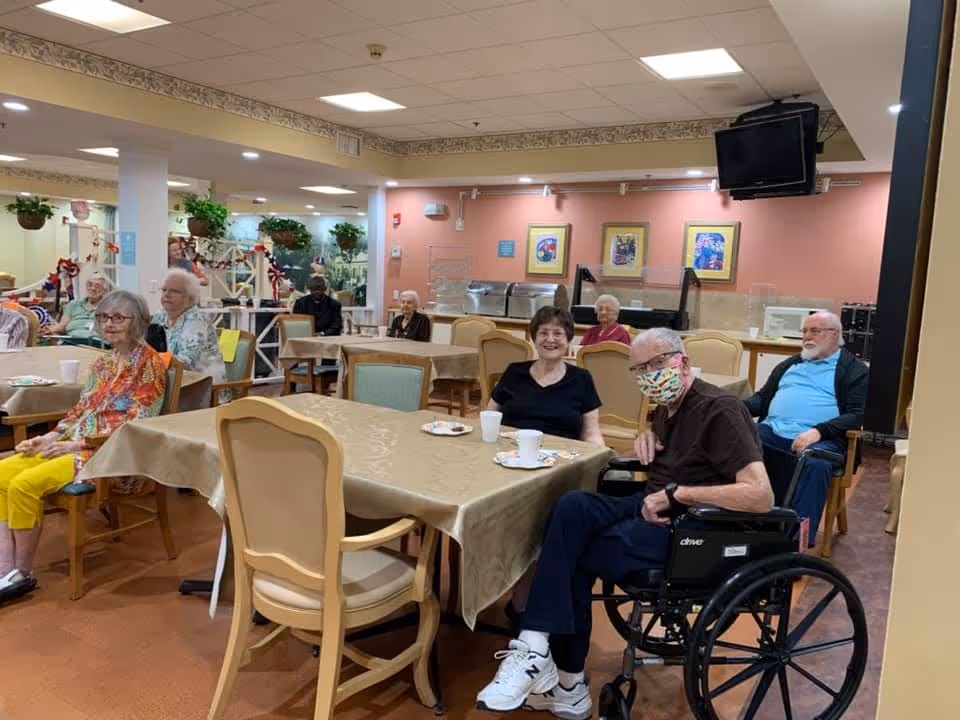 A group of elderly people sitting at tables in a senior living facility dining room. Some are seated in chairs while one person is in a wheelchair wearing a colorful face mask. The room has beige walls with framed artwork, a television mounted in the corner, and several tables covered with beige tablecloths. There are plants and decorations in the background.