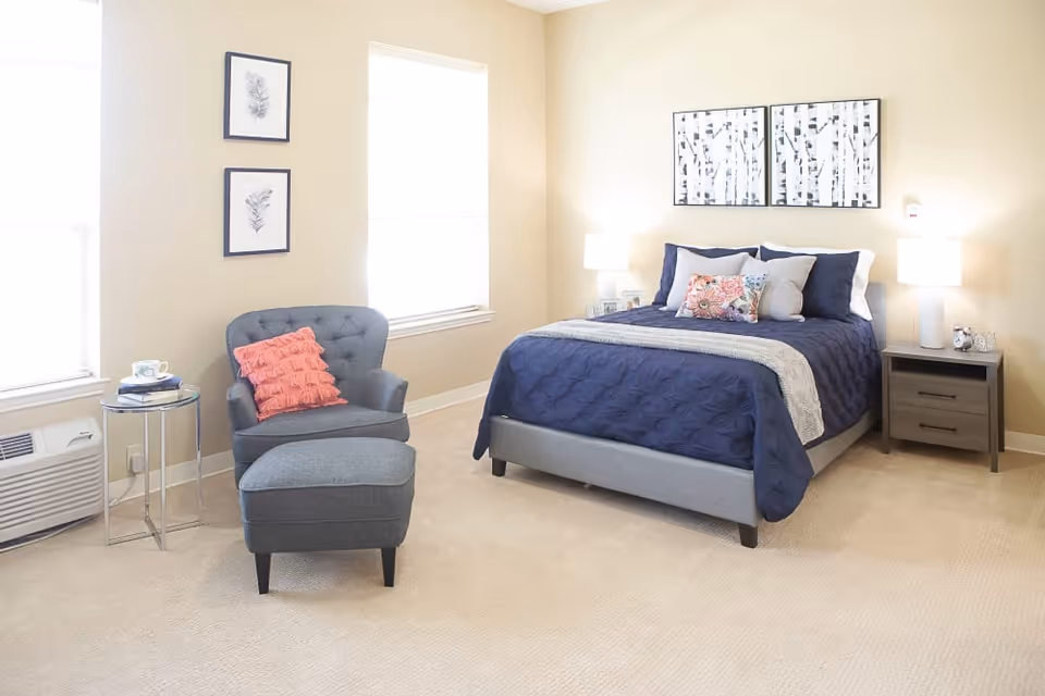 A bright bedroom with beige walls and carpet. The room features a bed with a navy blue quilt, multiple pillows, and a light gray throw blanket. On either side of the bed are matching nightstands with white lamps. To the left of the bed is a gray upholstered armchair with a matching ottoman and a coral-colored pillow. Next to the chair is a small round glass side table with a cup and books. Two framed botanical prints hang on the wall between two windows, and above the bed are two framed abstract art pieces.