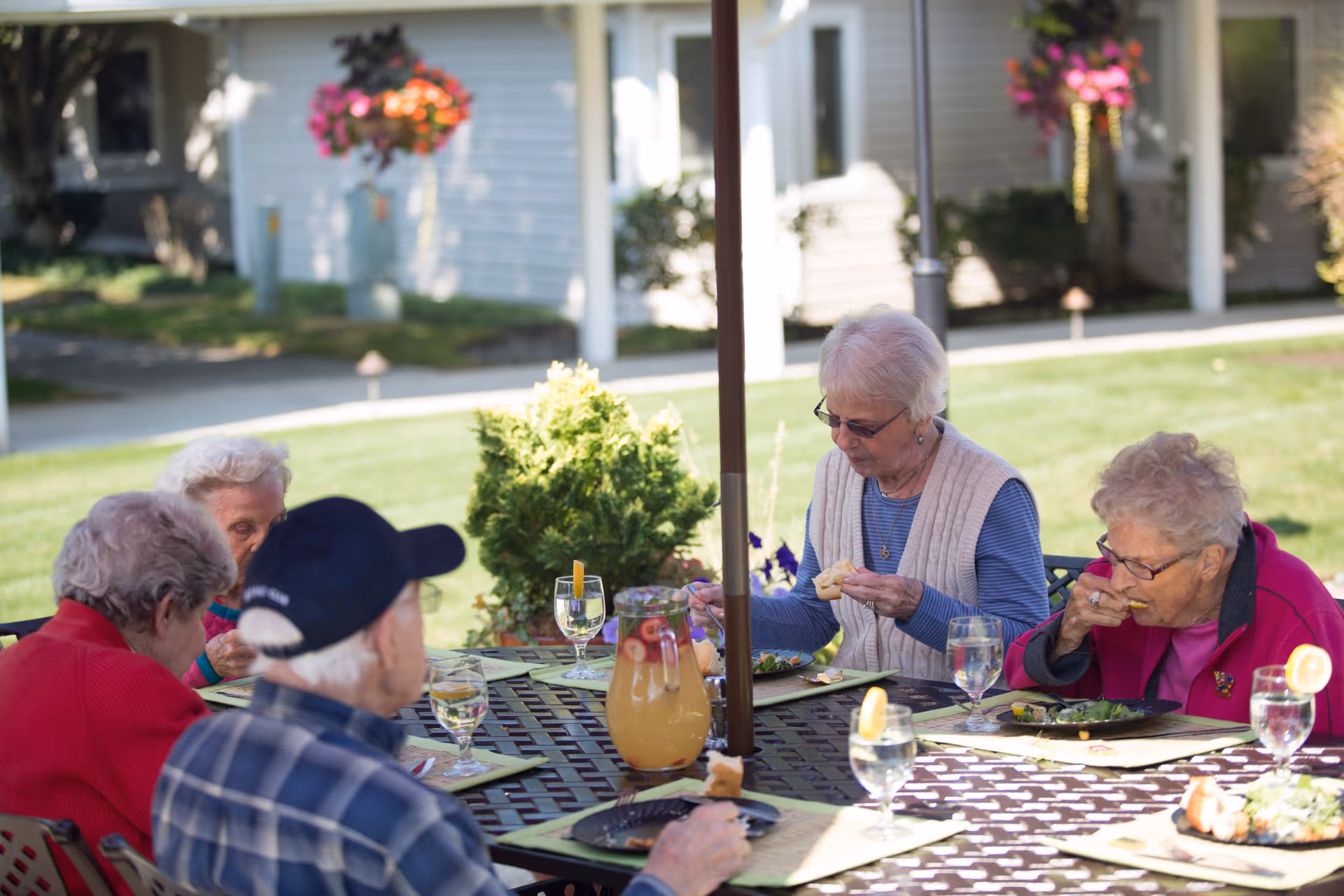 A group of elderly people sitting around a metal outdoor table under an umbrella, eating a meal together. There is a pitcher of fruit-infused water and glasses with lemon slices on the table. The background shows a green lawn, bushes, and a building with hanging flower baskets.