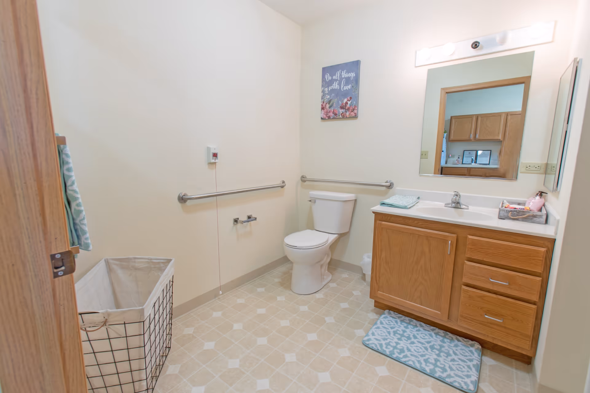 A clean and accessible bathroom with a toilet, wooden vanity with a sink, a mirror above the sink, grab bars on the walls, a laundry basket, and a decorative wall sign that reads 'Do all things with love'.