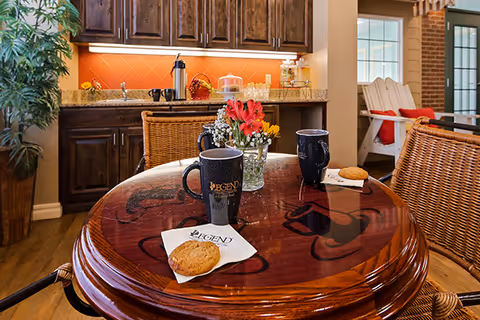 A cozy kitchen and dining area with a round wooden table featuring two black mugs, each on a napkin with the Legend logo, and two cookies. The background shows dark wooden cabinets, a countertop with a coffee pot, cups, and a small flower arrangement. There are wicker chairs around the table and a white wooden chair with a red cushion near a window.