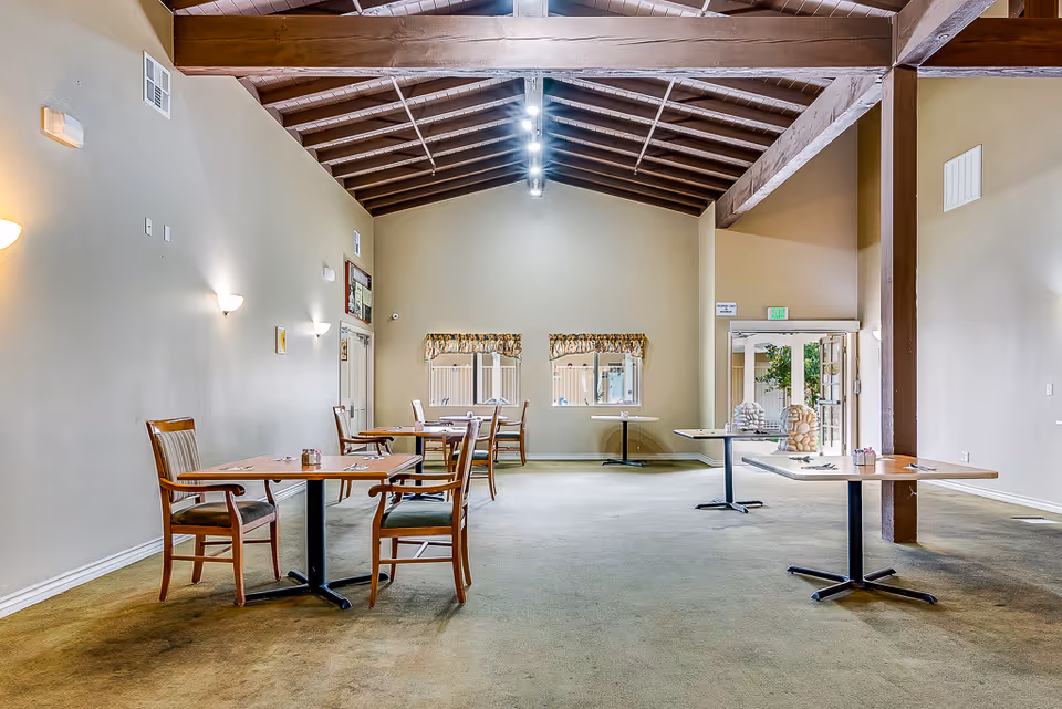A spacious dining area with several wooden tables and chairs arranged neatly. The room features a high ceiling with exposed wooden beams and track lighting. There are two windows with floral valances on the far wall and a glass door leading outside. The carpet is greenish, and the walls are painted beige with wall-mounted lights.