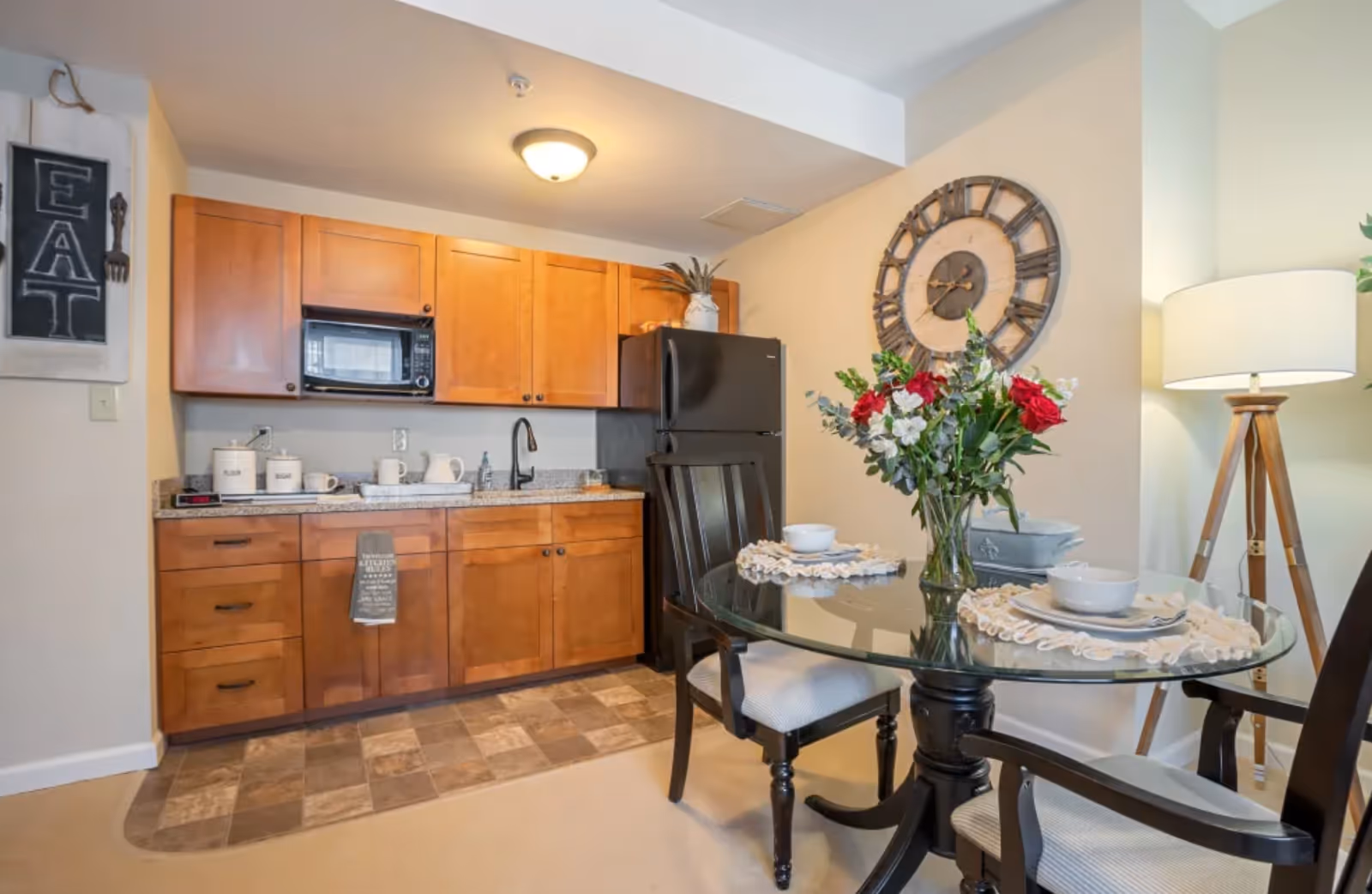 Small kitchenette with wooden cabinets and a black refrigerator beside a glass dining table set with flowers and place settings.